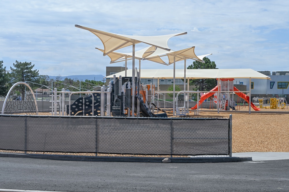 A modern, well-equipped playground area at what appears to be a school or community facility. The playground features multiple play structures, shade sails, and slides, all enclosed by a chain-link fence with a privacy screen. The area appears newly installed or recently renovated, with wood chip surfacing and no visible occupants.