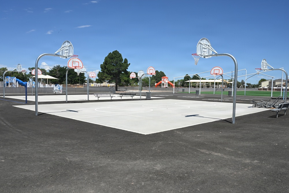 An outdoor public park basketball court complex with multiple hoops arranged around a light-colored concrete playing surface. The facility appears newly constructed or recently renovated, surrounded by asphalt and featuring additional park amenities in the background. The area is unoccupied and photographed under clear blue skies during daytime.