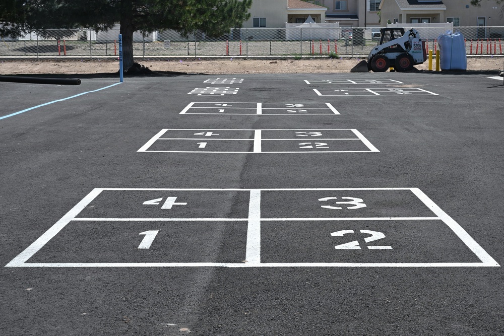 A freshly paved asphalt surface features multiple sets of shuffleboard court markings painted in white, receding into the distance in perspective. The courts are newly painted with numbered zones (1, 2, 3, 4) and appear to be part of a recreational area under construction or renovation. A skid-steer loader (Bobcat) and construction materials are visible in the background near a chain-link fence.