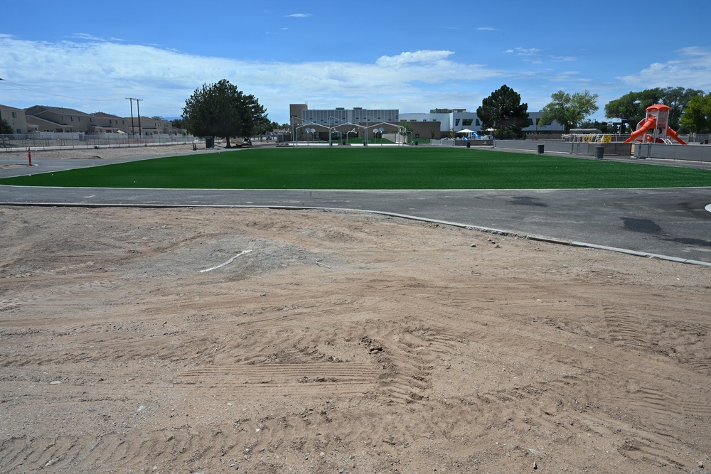 The image shows a school or park facility under construction or renovation, featuring a newly installed bright green artificial turf field surrounded by bare compacted dirt. A playground with an orange slide is visible on the right side, along with basketball hoops and covered structures in the background. The surrounding area appears to be in an arid, desert-like environment with residential and institutional buildings nearby.