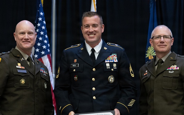 Three U.S. Army personnel are posed together at a formal military ceremony. The central figure, identified by his name tag as 'REEL,' is holding a diploma/certificate from the 120th Regiment Regional Training Institute made out to 'Coire R. Reel.' The two flanking individuals wear Army Service Uniforms in olive green, while the central figure wears the Army Service Uniform (blue dress uniform).
