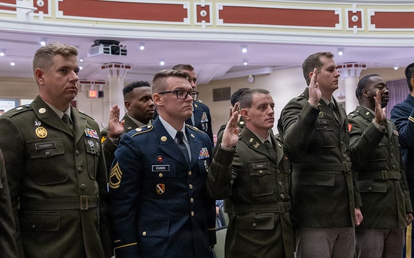 A group of military personnel in various U.S. Army uniforms are standing in a formal indoor ceremony, raising their right hands as if taking an oath. The setting appears to be an official military commissioning, promotion, or re-enlistment ceremony held in a formal hall or auditorium. Both male and female service members are present, wearing dress and service uniforms.