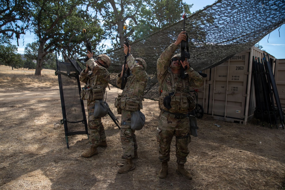 Three soldiers in MultiCam/OCP camouflage uniforms are holding rifles overhead, likely performing a weapons clearing or inspection procedure in an outdoor training area. They are positioned beneath a camouflage netting canopy near a tan military shipping container. The setting appears to be a dry, wooded training installation consistent with a California military base.