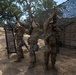 Three soldiers in MultiCam/OCP camouflage uniforms are holding rifles overhead, likely performing a weapons clearing or inspection procedure in an outdoor training area. They are positioned beneath a camouflage netting canopy near a tan military shipping container. The setting appears to be a dry, wooded training installation consistent with a California military base.