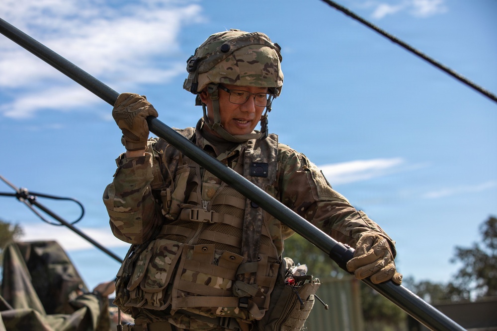 A soldier in full combat gear is handling a long metal pole or antenna mast in an outdoor field environment. The individual is wearing MultiCam/OCP camouflage uniform, tactical vest, combat helmet, glasses, and tactical gloves. The setting appears to be a military field exercise or communications setup operation.
