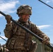 A soldier in full combat gear is handling a long metal pole or antenna mast in an outdoor field environment. The individual is wearing MultiCam/OCP camouflage uniform, tactical vest, combat helmet, glasses, and tactical gloves. The setting appears to be a military field exercise or communications setup operation.