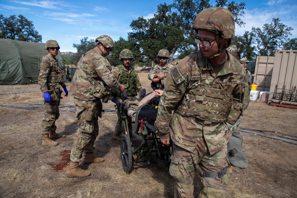 U.S. Army soldiers are conducting a medical training exercise at what appears to be a field camp or training installation. Personnel are transporting a simulated casualty on a wheeled litter/stretcher, with at least one soldier wearing blue medical gloves indicating a medical role. The scene includes multinational participants, as evidenced by one individual wearing a different camouflage pattern uniform.