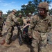 U.S. Army soldiers are conducting a medical training exercise at what appears to be a field camp or training installation. Personnel are transporting a simulated casualty on a wheeled litter/stretcher, with at least one soldier wearing blue medical gloves indicating a medical role. The scene includes multinational participants, as evidenced by one individual wearing a different camouflage pattern uniform.