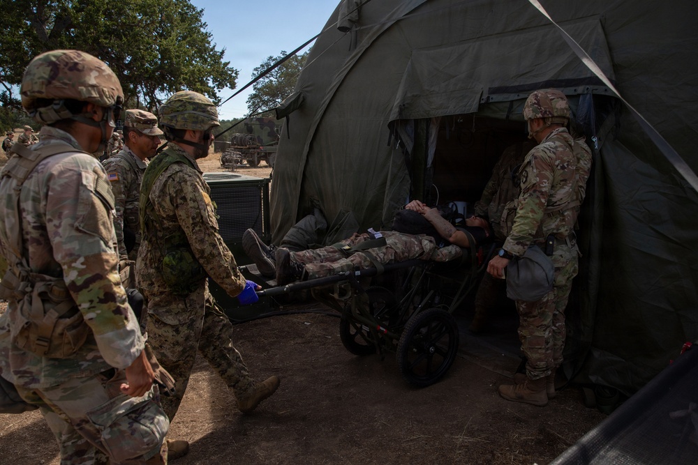 Military personnel in camouflage uniforms are transporting a casualty on a wheeled litter/stretcher into a large olive-drab military medical tent. The scene appears to be a field medical exercise or training scenario, with multiple soldiers assisting in the patient transfer. The setting is an outdoor military field environment with trees and military vehicles visible in the background.