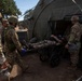 Military personnel in camouflage uniforms are transporting a casualty on a wheeled litter/stretcher into a large olive-drab military medical tent. The scene appears to be a field medical exercise or training scenario, with multiple soldiers assisting in the patient transfer. The setting is an outdoor military field environment with trees and military vehicles visible in the background.