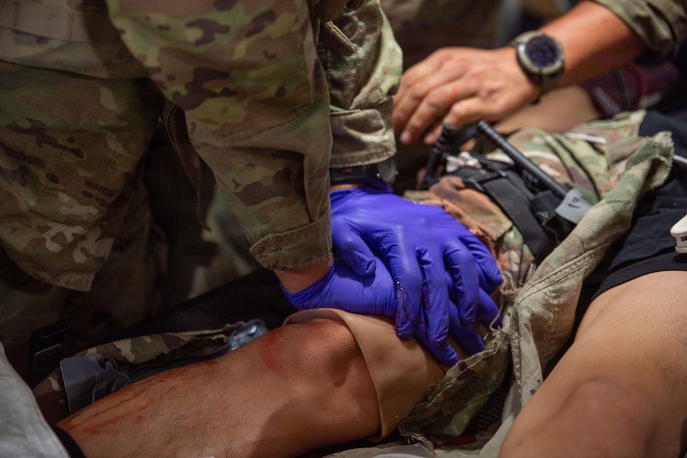 A military medic wearing blue nitrile gloves is providing tactical combat casualty care (TCCC) to a patient in military uniform. The medic appears to be applying pressure or treating a wound in the abdominal/hip area of the casualty. A second person wearing a watch is visible assisting in the background, suggesting this may be a training exercise or real field medical response.