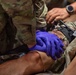 A military medic wearing blue nitrile gloves is providing tactical combat casualty care (TCCC) to a patient in military uniform. The medic appears to be applying pressure or treating a wound in the abdominal/hip area of the casualty. A second person wearing a watch is visible assisting in the background, suggesting this may be a training exercise or real field medical response.