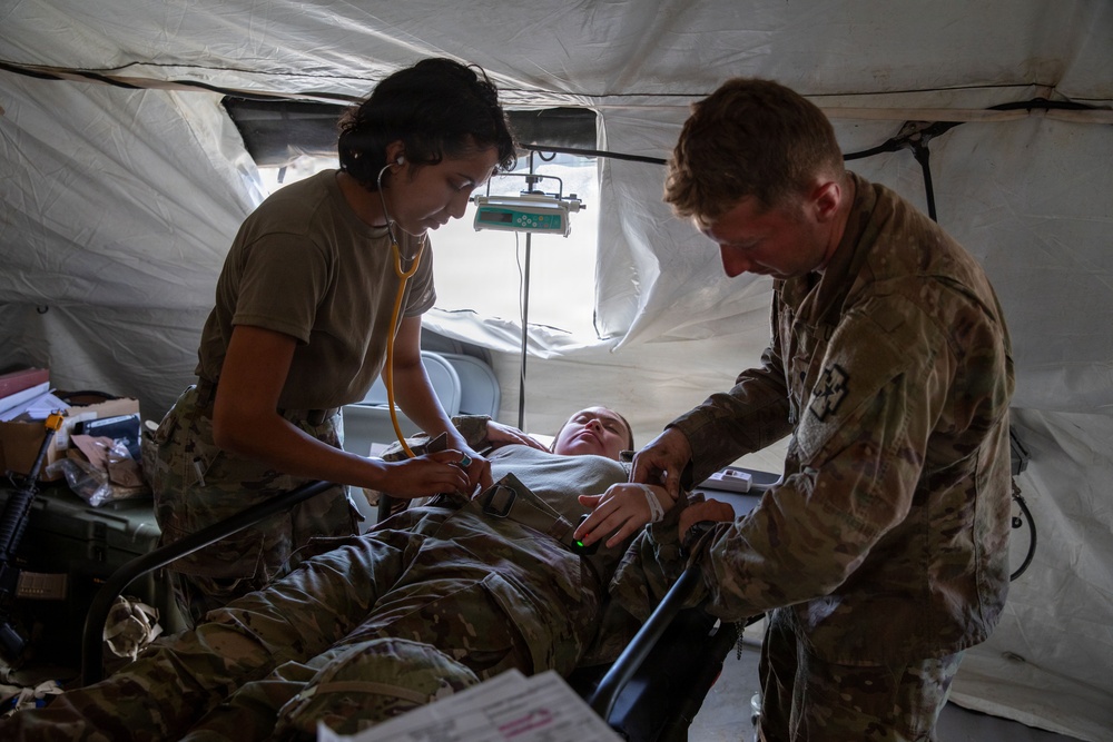 Two military medical personnel in camouflage uniforms are treating a patient lying on a field medical cot inside a military tent. One provider is using a stethoscope while the other appears to be checking vitals or administering treatment. The setting appears to be a forward operating base or field medical station.