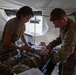 Two military medical personnel in camouflage uniforms are treating a patient lying on a field medical cot inside a military tent. One provider is using a stethoscope while the other appears to be checking vitals or administering treatment. The setting appears to be a forward operating base or field medical station.