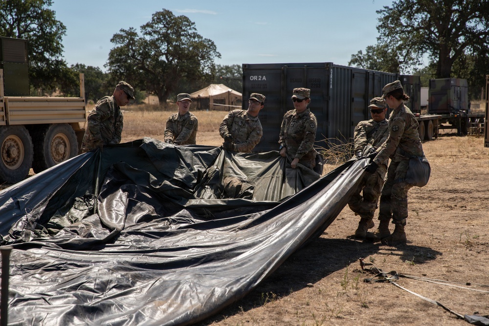A group of approximately six military personnel in camouflage uniforms are working together to fold or deploy a large black tarp or tent material in an outdoor field setting. The scene appears to be a military base camp or training area, with shipping containers, military vehicles, and a tent visible in the background. The environment suggests a dry, warm climate with sparse vegetation and oak trees.