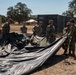 A group of approximately six military personnel in camouflage uniforms are working together to fold or deploy a large black tarp or tent material in an outdoor field setting. The scene appears to be a military base camp or training area, with shipping containers, military vehicles, and a tent visible in the background. The environment suggests a dry, warm climate with sparse vegetation and oak trees.