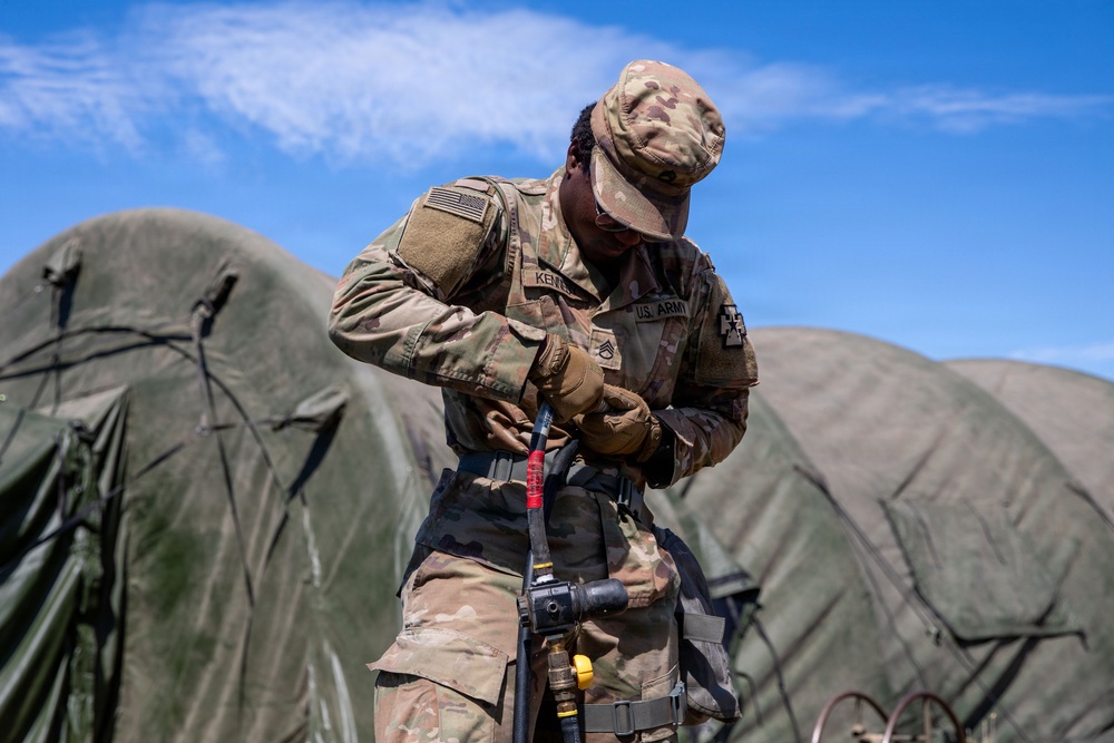 A U.S. Army soldier in MultiCam/OCP camouflage uniform is working with fuel or fluid transfer equipment, likely a fuel nozzle or hose assembly, at what appears to be a forward operating base or field camp. The soldier, whose name tape reads 'KENNE' (partially visible) and wears a 2nd Infantry Division patch on the right shoulder, is focused on connecting or inspecting the equipment. Green military inflatable or dome-style tents are visible in the background.