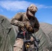 A U.S. Army soldier in MultiCam/OCP camouflage uniform is working with fuel or fluid transfer equipment, likely a fuel nozzle or hose assembly, at what appears to be a forward operating base or field camp. The soldier, whose name tape reads 'KENNE' (partially visible) and wears a 2nd Infantry Division patch on the right shoulder, is focused on connecting or inspecting the equipment. Green military inflatable or dome-style tents are visible in the background.