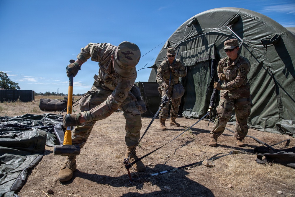 Three soldiers in U.S. Army multicam/OCP uniforms are setting up or securing a large military field tent in an arid, open terrain environment. The foreground soldier is using a yellow-handled sledgehammer to drive a ground stake, while two other soldiers observe and assist nearby. The setting appears to be a field training exercise or forward operating base establishment.