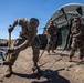 Three soldiers in U.S. Army multicam/OCP uniforms are setting up or securing a large military field tent in an arid, open terrain environment. The foreground soldier is using a yellow-handled sledgehammer to drive a ground stake, while two other soldiers observe and assist nearby. The setting appears to be a field training exercise or forward operating base establishment.