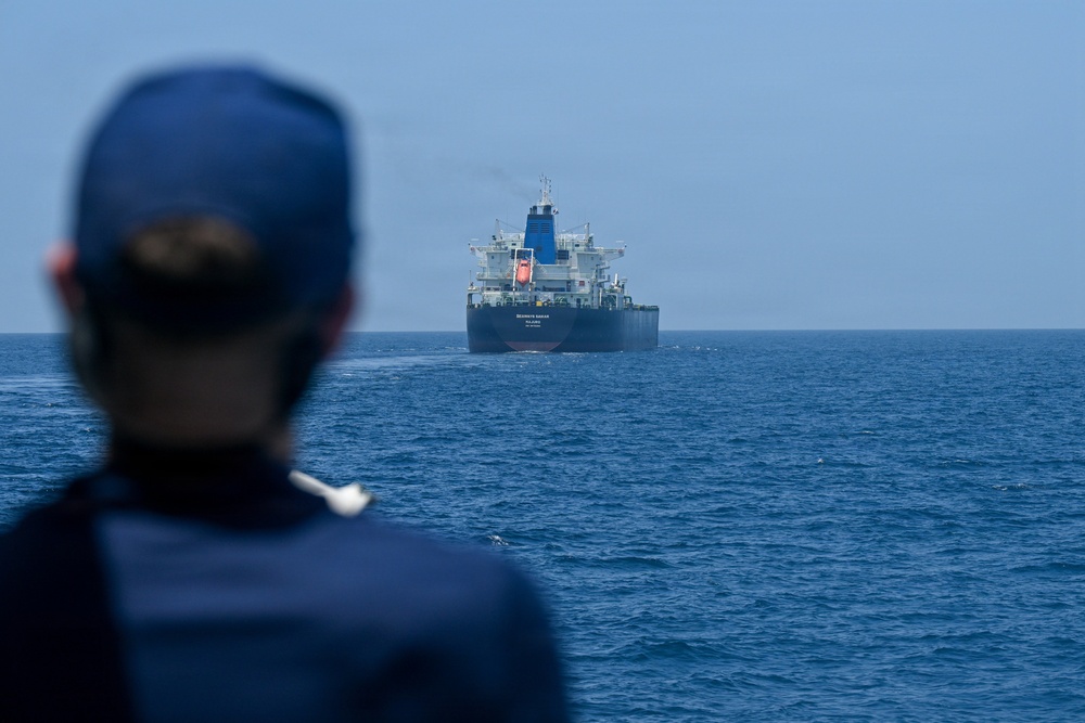 A person wearing a dark blue uniform and cap is observed from behind, watching a large dark-hulled vessel — appearing to be an oil tanker or bulk carrier — anchored or underway in open blue waters. The ship displays white superstructure and appears to have text on its hull, though it is not fully legible at this distance. The scene suggests a maritime observation or monitoring activity.