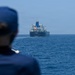 A person wearing a dark blue uniform and cap is observed from behind, watching a large dark-hulled vessel — appearing to be an oil tanker or bulk carrier — anchored or underway in open blue waters. The ship displays white superstructure and appears to have text on its hull, though it is not fully legible at this distance. The scene suggests a maritime observation or monitoring activity.