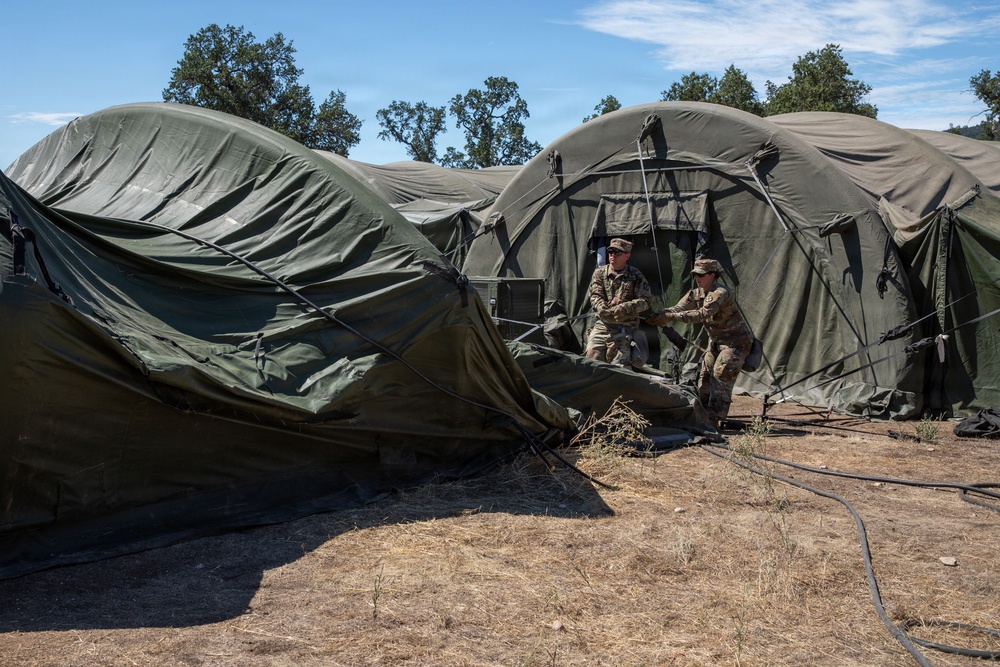 Two military personnel in camouflage uniforms are working with large olive-drab military field tents in an outdoor setting. The soldiers appear to be either setting up or breaking down a multi-section modular tent system. The environment is dry and arid with sparse vegetation and deciduous trees visible in the background.