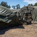 Two military personnel in camouflage uniforms are working with large olive-drab military field tents in an outdoor setting. The soldiers appear to be either setting up or breaking down a multi-section modular tent system. The environment is dry and arid with sparse vegetation and deciduous trees visible in the background.