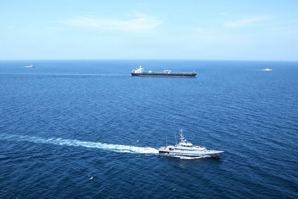 An aerial photograph shows a naval or coast guard patrol vessel in the foreground escorting or monitoring a large dark-hulled commercial tanker in open blue ocean waters. Two additional smaller vessels are visible in the far background on either side of the tanker. The scene suggests a maritime escort, interception, or patrol operation in calm sea conditions.