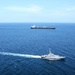 An aerial photograph shows a naval or coast guard patrol vessel in the foreground escorting or monitoring a large dark-hulled commercial tanker in open blue ocean waters. Two additional smaller vessels are visible in the far background on either side of the tanker. The scene suggests a maritime escort, interception, or patrol operation in calm sea conditions.