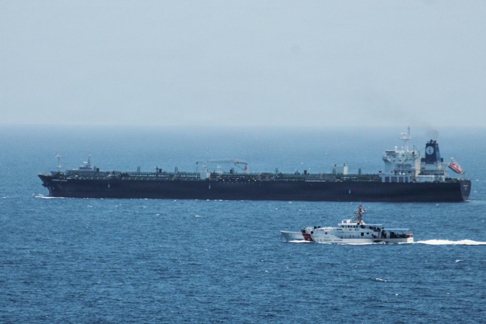 The image shows a large black oil tanker vessel at sea accompanied by a smaller white U.S. Coast Guard patrol boat. The Coast Guard vessel appears to be escorting or intercepting the tanker in open ocean waters. The scene suggests a maritime law enforcement or escort operation.