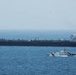 The image shows a large black oil tanker vessel at sea accompanied by a smaller white U.S. Coast Guard patrol boat. The Coast Guard vessel appears to be escorting or intercepting the tanker in open ocean waters. The scene suggests a maritime law enforcement or escort operation.