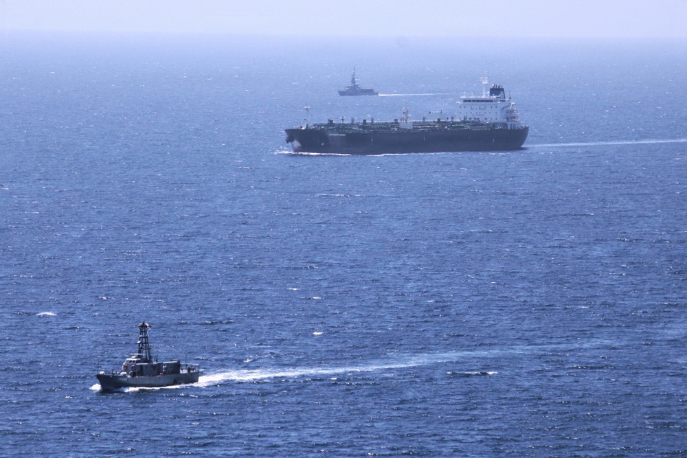The image shows three vessels at sea under hazy atmospheric conditions. A large dark-hulled cargo or oil tanker occupies the middle ground, flanked by two smaller patrol or naval vessels. The scene suggests a maritime escort or monitoring operation.