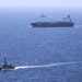 The image shows three vessels at sea under hazy atmospheric conditions. A large dark-hulled cargo or oil tanker occupies the middle ground, flanked by two smaller patrol or naval vessels. The scene suggests a maritime escort or monitoring operation.