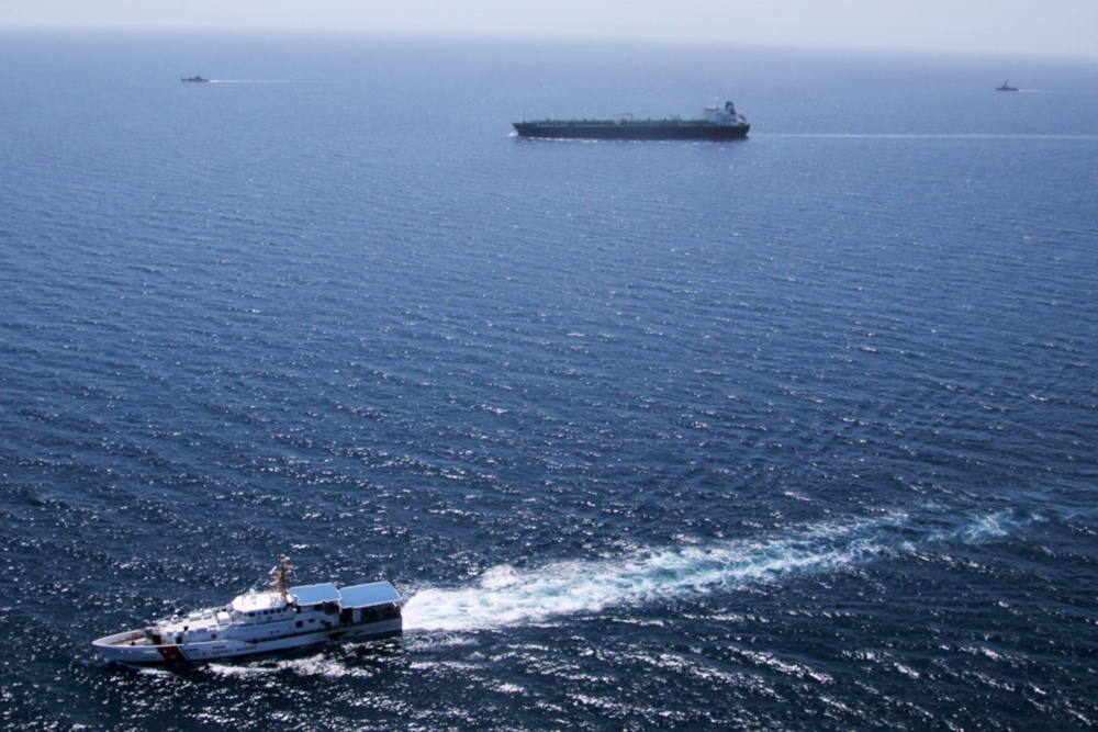 An aerial photograph showing multiple vessels on open ocean waters. A white patrol/coast guard vessel is prominently visible in the foreground creating a wake, with a large dark-hulled cargo or tanker ship in the middle distance, and two smaller vessels visible on the far horizon. The scene suggests a maritime escort, intercept, or monitoring operation.