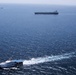 An aerial photograph showing multiple vessels on open ocean waters. A white patrol/coast guard vessel is prominently visible in the foreground creating a wake, with a large dark-hulled cargo or tanker ship in the middle distance, and two smaller vessels visible on the far horizon. The scene suggests a maritime escort, intercept, or monitoring operation.