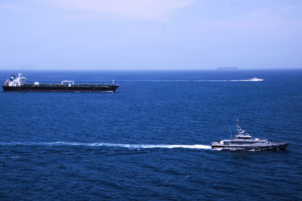 The image shows an open ocean scene with multiple vessels visible. A large dark-hulled tanker ship is positioned in the left background, while a smaller gray patrol or coast guard vessel is moving at speed in the foreground right, leaving a visible wake. A third smaller fast-moving vessel is visible in the mid-distance background.