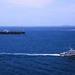 The image shows an open ocean scene with multiple vessels visible. A large dark-hulled tanker ship is positioned in the left background, while a smaller gray patrol or coast guard vessel is moving at speed in the foreground right, leaving a visible wake. A third smaller fast-moving vessel is visible in the mid-distance background.