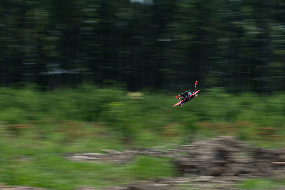 A small FPV racing drone is captured mid-flight against a blurred green background of trees and grass, indicating high speed movement. The panning camera technique has kept the drone relatively sharp while the background shows significant motion blur. The drone appears to be red and black in color and is tilted at an angle suggesting aggressive maneuvering.