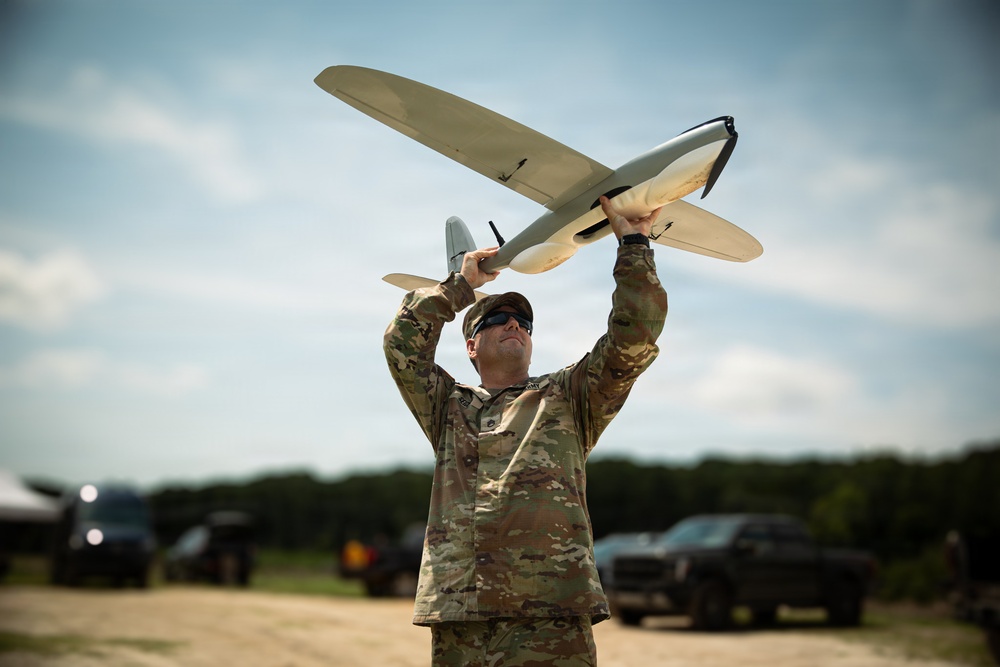 A military service member in camouflage uniform is preparing to hand-launch a small fixed-wing unmanned aerial vehicle (UAV) overhead. The setting appears to be an outdoor field or staging area with vehicles and tree line visible in the background. The image captures a training or operational deployment of a tactical reconnaissance drone.