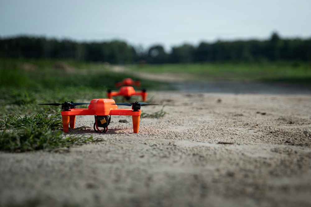 Two bright orange quadcopter drones are positioned on a sandy dirt path in a rural outdoor setting. The foreground drone is in sharp focus, revealing four black propellers, orange landing legs, and a small yellow component underneath the body. A second identical drone is visible in the background, slightly out of focus along the same path.