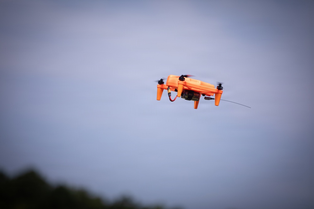 An orange quadcopter drone is captured mid-flight against an overcast blue-grey sky. The drone appears to be carrying or trailing a thin wire or antenna extending from its underside. Treetops are faintly visible in the lower left corner of the frame.