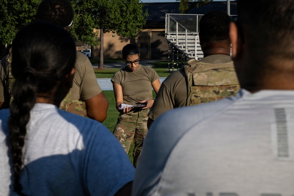 A female military service member in OCP (Operational Camouflage Pattern) uniform is addressing a group of individuals outdoors on what appears to be a military installation. She is holding papers or a clipboard and appears to be conducting a briefing or roll call. The setting includes a grassy field with bleachers and a chain-link fence structure visible in the background.