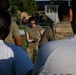 A female military service member in OCP (Operational Camouflage Pattern) uniform is addressing a group of individuals outdoors on what appears to be a military installation. She is holding papers or a clipboard and appears to be conducting a briefing or roll call. The setting includes a grassy field with bleachers and a chain-link fence structure visible in the background.