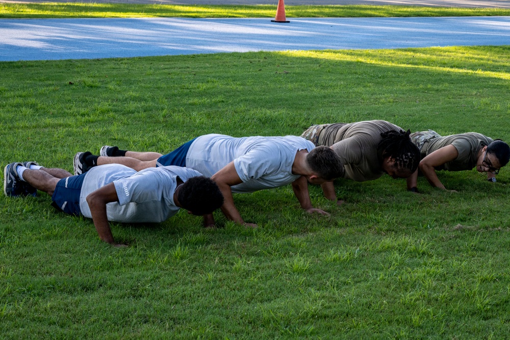 A group of individuals are performing push-ups in a line on a grassy field. Some participants are wearing military-style camouflage clothing while others are in civilian athletic wear, suggesting a mixed military and civilian fitness training session. The setting appears to be an outdoor area near a road, indicated by the visible traffic cone in the background.