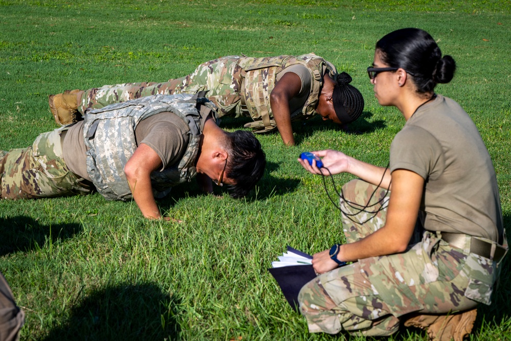 Two soldiers in camouflage uniforms and body armor are performing push-ups on a grass field while a third soldier kneels nearby holding a stopwatch and clipboard, apparently serving as a grader or evaluator. The scene appears to be a military physical fitness test or assessment conducted outdoors during daylight hours. The evaluator is closely observing the soldiers' form and tracking their performance.