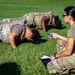 Two soldiers in camouflage uniforms and body armor are performing push-ups on a grass field while a third soldier kneels nearby holding a stopwatch and clipboard, apparently serving as a grader or evaluator. The scene appears to be a military physical fitness test or assessment conducted outdoors during daylight hours. The evaluator is closely observing the soldiers' form and tracking their performance.