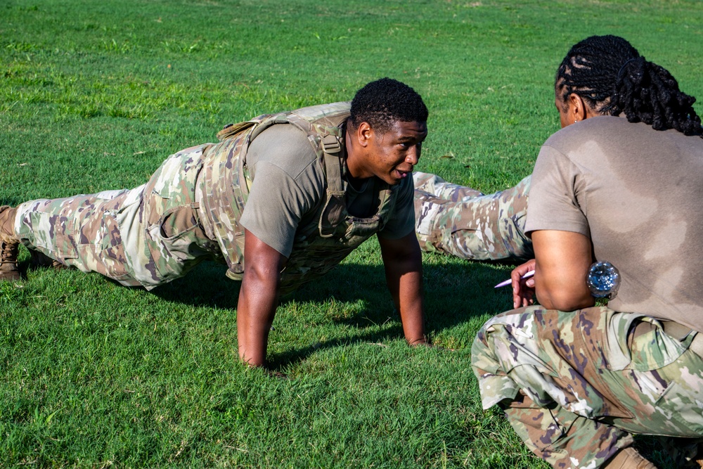 A soldier in OCP (Operational Camouflage Pattern) uniform is performing push-ups on a grass field while a second soldier, appearing to be a grader or evaluator, crouches nearby holding a pen and paper. This appears to be a military physical fitness test or assessment. The setting is an outdoor grassy area in daylight.