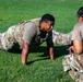 A soldier in OCP (Operational Camouflage Pattern) uniform is performing push-ups on a grass field while a second soldier, appearing to be a grader or evaluator, crouches nearby holding a pen and paper. This appears to be a military physical fitness test or assessment. The setting is an outdoor grassy area in daylight.
