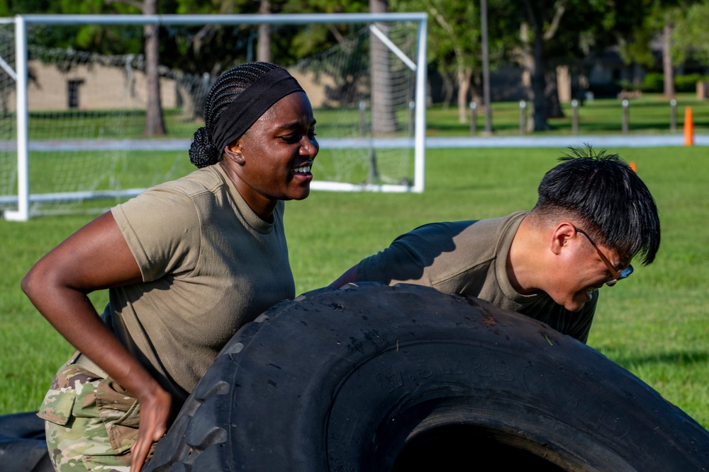Two individuals in military-style olive/camouflage uniforms are performing a tire flip exercise on a grass field. A woman with braided hair and a black headband is visible on the left, while a man wearing glasses is pushing from the right side. The activity appears to be part of a physical training or fitness challenge event.